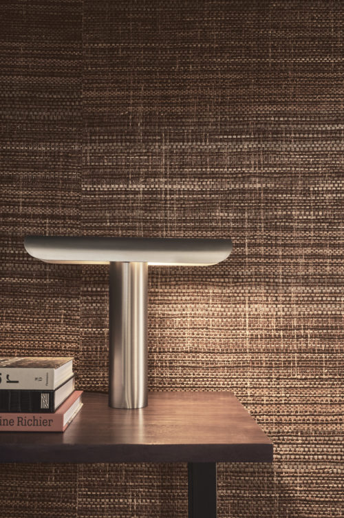 Closeup of a wooden side table placed against a wall, with a sleek minimalist metal table lamp and a pile of books on top of it. As a backdrop, Omexco’s handmade natural wallcoverings collection “Intuition”. This wallcovering design reveals itself in shades of brown that intertwine harmoniously, punctuated by the skillful weaving of various materials such as bakbak, bacnoc, and paper yarn.