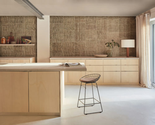 Captivating kitchen shot featuring Omexco Intuition wallcoverings, meticulously crafted from burlap with an elegant patchwork design and exquisite stitching in a natural brown color. The modern open kitchen has a light grey cast floor and features a sleek design with light wood cupboards with drawers. In the center of the image, a metal sleek barstool stands next to the kitchen island, providing a stylish and functional seating option. On the right side of the image, a large window adorned with long linen curtains lets in natural light, creating a bright and airy atmosphere in the kitchen.