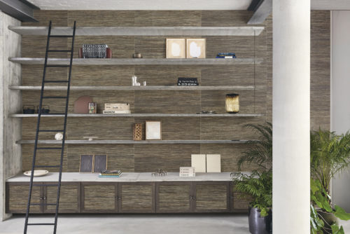 An airy loft-inspired living room featuring a concrete open bookcase with floating wall shelves, adorned with books and curated decorative elements. A sleek black ladder leans against the left side, providing access to the upper shelves. On the right, a beige column and indoor plants add a touch of natural elegance. The space is grounded by a grey cast floor, and the backdrop showcases Omexco's handmade natural Intuition wallcovering collection with a textured ramie weave in harmonious shades of brown and grey, creating a sophisticated and inviting ambiance.