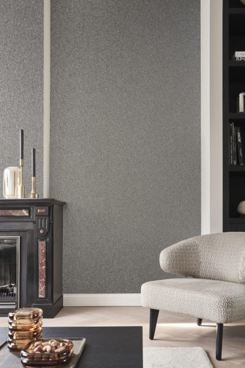 In this captivating decor photo, a stylish black fireplace takes center stage, complemented by a sleek black coffee table and a chic black-and-white armchair. The monochromatic theme is enhanced by a pristine white carpet, creating a modern and sophisticated ambiance. The wall showcases the elegance of Omexco's Archipelago collection, featuring embossed grey mica wallcoverings that contribute to the overall sophistication of the scene.