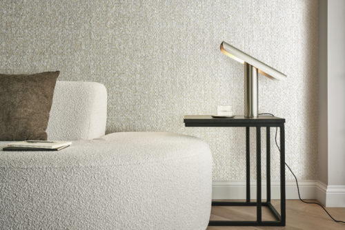 A close-up of a cozy living room featuring a beige bouclé fabric couch adorned with a brown pillow. The herringbone parquet floor complements the classic skirting boards, creating a timeless aesthetic. A black side table stands nearby, adorned with a minimalist golden table lamp. As a stunning backdrop, Omexco's capiz mother of pearl wallcoverings from the Archipelago collection showcase shimmering shades of beige, adding a touch of elegance and sophistication to the intimate space.