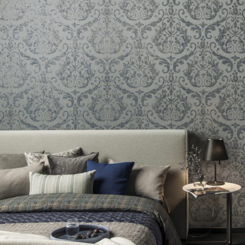 We zoom in on a tactile blue and grey bedroom. Soft pillows in different colours, plaids in different structures. Black bedside lamp. Vase with purple flowers.