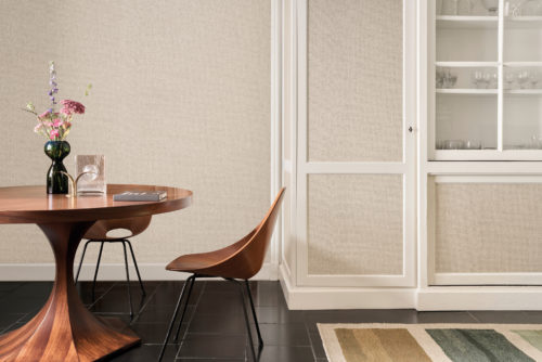 This elegant dining space highlights the OAS274 reference, titled Xico, from Omexco’s Oase collection. The wallpaper, crafted from plain paperweave, showcases a refined tone-on-tone design in warm, earthy hues. The subtle texture adds depth and tactility while maintaining a harmonious, natural aesthetic. The wallcovering extends beyond the walls, seamlessly integrating into the cabinetry, creating a cohesive and sophisticated look. The rich wood tones of the dining table and chair contrast beautifully against the soft, neutral backdrop, while the floral arrangement and decorative glassware add a refined touch to the setting.
