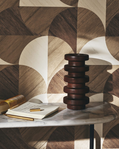 Close-up of geometric marquetry wallcoverings from Omexco's Imperialis collection, featuring shades of light brown, dark brown and beige. Inspired by lotus leaves, the design exudes grace and elegance. In the foreground, a marble side table holds an open notebook and a wooden decorative element. Soft daylight illuminates the wall, enhancing the intricate patterns and textures.