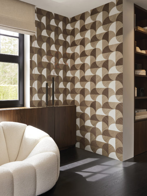 A cozy living room corner featuring a dark grey parquet floor, a white comfortable couch on the left and a window, providing natural light. On the right, built-in wall shelves display books. Adorning the wall is Omexco's marquetry collection Imperialis, showcasing geometric shapes in light brown, dark brown, and white, inspired by the graceful shape of lotus leaves.