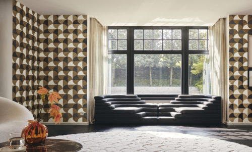 A spacious living room with a large window in the center, offering natural light. In front of the window sits a sleek black leather couch. On the left, a marble side table holds an orange glass vase. A large round white carpet contrasts against the dark grey parquet floor. Adorning the wall is Omexco's marquetry collection Imperialis, featuring geometric shapes in brown, black, and white, inspired by the graceful shape of lotus leaves.