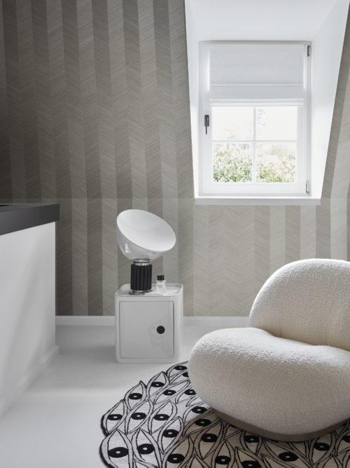 A serene room with neutral tones, featuring a sloped roof and a window. The floor is white with a light grey and black rug in an eye-catching pattern. A comfortable single seater upholstered in bouclé fabric sits atop the rug. Adjacent to it is a sleek white nightstand with a table lamp. On the wall, Omexco's marquetry collection Imperialis is displayed, showcasing a grey herringbone motif of fine wood veneer expertly hand-inlaid for elegant simplicity.