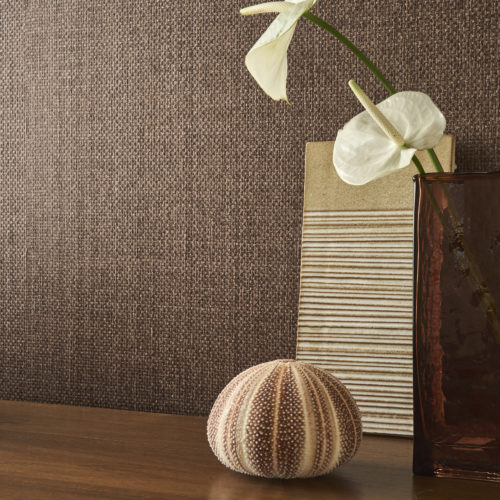 This elegant setting showcases the OAS291 reference, titled Cesta, from Omexco’s Oase collection. The wallcovering is crafted from finely woven paper fibres, dyed with gentle irregularity to capture the organic nuances of natural materials. The deep, earthy hues radiate warmth and authenticity, reinforcing the tactile appeal of the textured surface. A composition of organic elements, including a ceramic relief panel, a smoked glass vase, and a delicately placed sea urchin shell, enhances the natural aesthetic. The scene is completed by a dark wooden surface that adds richness and depth, beautifully complementing the wallpaper’s refined simplicity.
