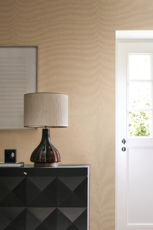 This refined interior showcases the OAS264 reference, titled Selva, from Omexco’s Oase collection. The wallpaper, composed of delicate paperweave, subtly highlights the organic beauty of its material. Its fine texture and soft, earthy hue bring warmth and sophistication to the space, creating a natural and inviting atmosphere. The elegant contrast between the dark geometric-patterned sideboard and the light wallcovering enhances the room’s visual balance, while the sculptural glass lamp and abstract artwork add a touch of modern artistry. Natural light filtering through the door gently accentuates the wallpaper’s texture, reinforcing the harmonious ambiance.