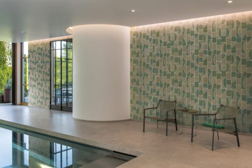 Image of a serene indoor pool area featuring two armchairs, a khaki round end table, a white rounded wall on the left and a green, geometric wallpaper on the right. This graphic pattern in light green and dark green is a playful pop of pattern which adds visual interest and modernity to the space. The mosaic of contrasting geometric shapes is made of cork and belongs to Omexco’s wallcovering collection “Casca”. The color palette and design elements create a peaceful and refreshing atmosphere that is perfect for relaxation.