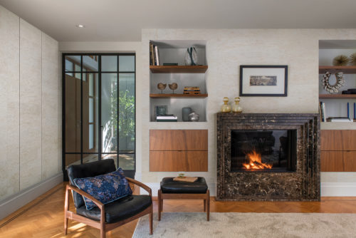 An image of a bright living room with a textured wallcovering in off-white featuring a subtle check pattern. These natural wallcoverings by Omexco have a leather-like look, but are actually made of Mulberry bark. The floor is covered with herringbone wood planks that add a touch of elegance and depth to the space. A wood and leather armchair with a matching footstool look inviting and relaxating. A built-in bookshelf with various objects and books as well as a burning fireplace are visible in the background. The interior door made of glass and black steel can be seen on the left of the bookshelf. The overall aesthetic is a harmonious blend of natural textures and sleek design elements, creating a cozy and inviting atmosphere.