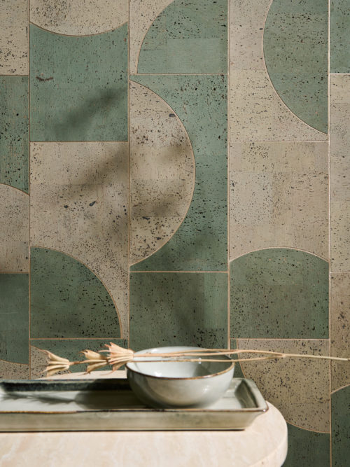 An image of a green, geometric wallpaper. The graphic pattern in light green and dark green is a playful pop of pattern which adds visual interest and modernity to the space. The mosaic of contrasting geometric shapes is made of cork and belongs to Omexco’s wallcovering collection “Casca”. In the foreground we see a wooden round side table with a light blue ceramic plate and small bowl placed on top of it.