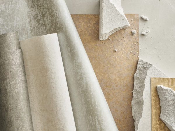 English:  An assortment of printed non-woven wallcoverings from Omexco's "Edition" collection, displayed in overlapping rolls. The collection features structured materials with plaster and patina effects, exuding a subtle hint of Bauhaus aesthetics. Pieces of textured plaster and patina materials are scattered around, creating a harmonious and luxurious display. The designs convey purity and serenity, making this collection a reference for architects and interior designers who value high-end quality and minimalist refinement.