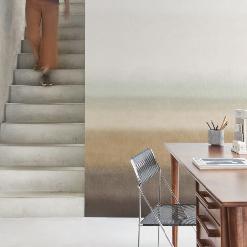 A person wearing a blue top and brown pants is descending a minimalist concrete staircase on the left side of the image. The background wall features Omexco’s printed non-woven “Edition” collection, a timeless and soft gradient design transitioning from from beige, light green and light brown to dark brown. On the right, there is a modern wooden desk with a few items, including a magazine, a cup with pencils, and a small decorative object. A sleek metal chair is positioned in front of the desk. The floor is light-colored, complementing the overall modern and minimalist aesthetic of the space.