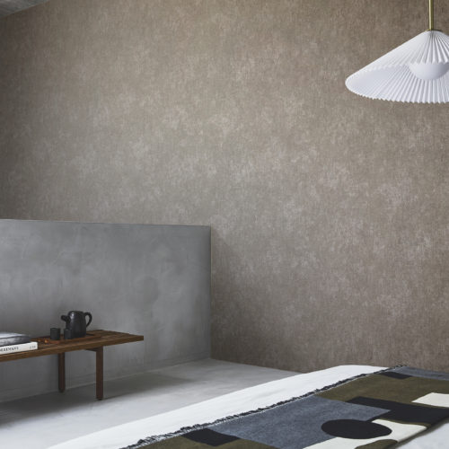 A room with a minimalist aesthetic. The back wall features a textured chocolate brown printed non-woven wallcovering from Omexco’s Edition collection. In the center, a hanging lamp with a white pleated lampshade is attached to the ceiling. To the left, a wooden bench holding books and a black teapot is placed in front of a smooth concrete wall. A bed is partially visible in the foreground, covered with a blanket featuring geometric patterns in black, gray, olive green, and beige. The floor is light-colored, complementing the modern and clean look of the room.