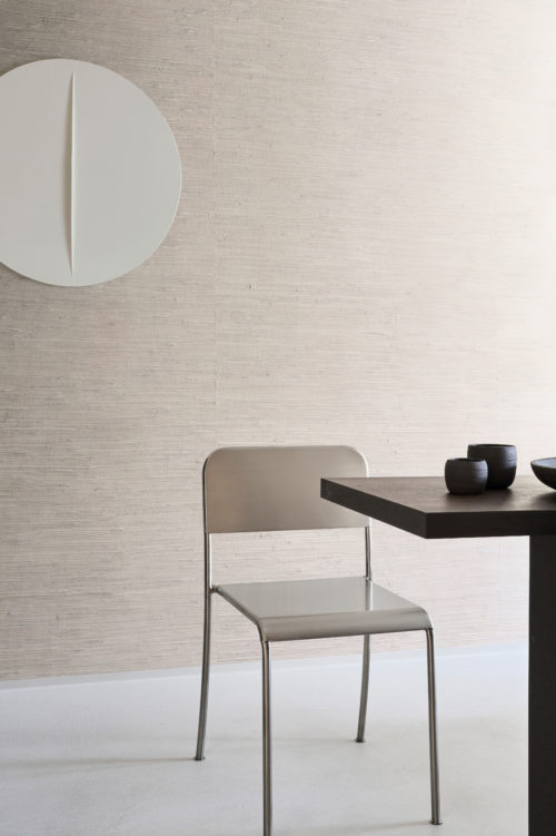 A minimalist dining area featuring Omexco's Pure collection, reference PU231, known as Osmos. This refined wallpaper showcases a delicate weave of natural waterlily fibres, gracefully finished with a soft layer of white paint. The understated design brings a sense of calm and sophistication to the space. A sleek metallic chair pairs harmoniously with a modern dark-stained dining table, complemented by minimalist ceramic bowls in muted tones. On the wall, a sculptural white art piece adds an abstract element, balancing the natural textures of the wallpaper. This setting seamlessly blends contemporary aesthetics with the organic beauty of natural materials.