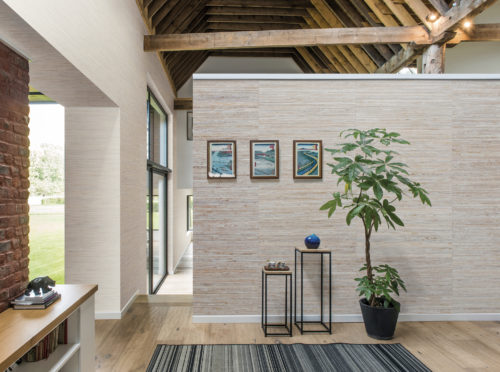 Bright room with a high ceiling. Lots of wood in the room. On the walls Shades of Pale wallcoverings in jute and tropical leaves. On the central wall 3 artworks water themed and two pedestalls with vases.