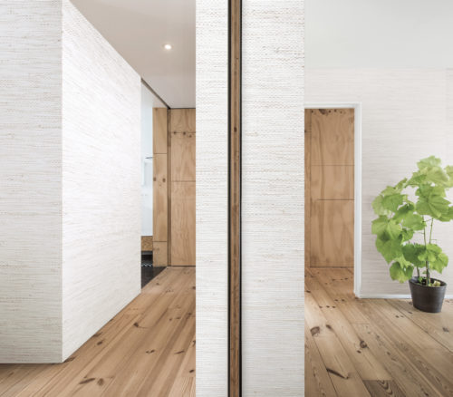 Room with wooden floors. The room is devided in two by a wall. On both sides of he wall bacnoc and paper weave wallcovering. It gives the bland room a lot of life. On the far right wall:  jute weave wallcovering.