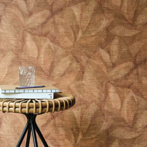 This inviting interior highlights Omexco’s OAS202 reference, Hierbas, from the Oase collection. The wallcovering features a delicate leaf motif, printed onto natural raffia, evoking an organic and warm atmosphere. The earthy terracotta tones of the wall harmonize with the rattan and metal side table, creating a setting that feels both rustic and refined. A stack of books, a glass of water, and a pen placed atop the table enhance the relaxed yet sophisticated ambiance, making this space ideal for moments of calm and contemplation.