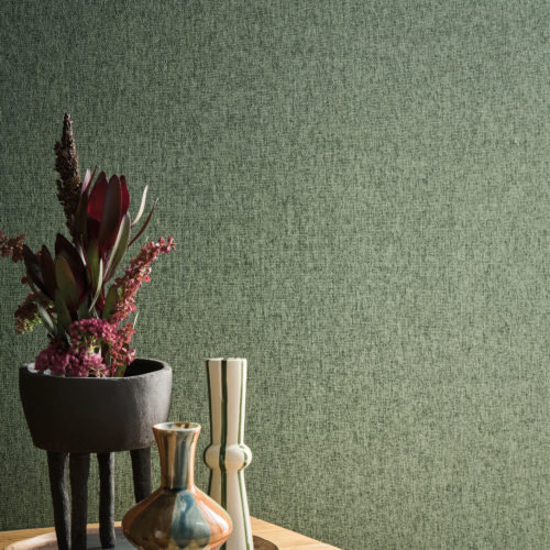 This refined interior highlights Omexco’s OAS272 reference, Xico, from the Oase collection. The wallcovering features a subtle paperweave texture in a tone-on-tone, earthy green hue, creating a natural and harmonious ambiance. The organic quality of the design is complemented by a wooden console, adorned with an eclectic mix of handcrafted ceramic vases and a sculptural black planter filled with lush, deep-hued florals. The interplay of textures and natural materials enhances the warm and inviting atmosphere, emphasizing the timeless beauty of artisanal craftsmanship.