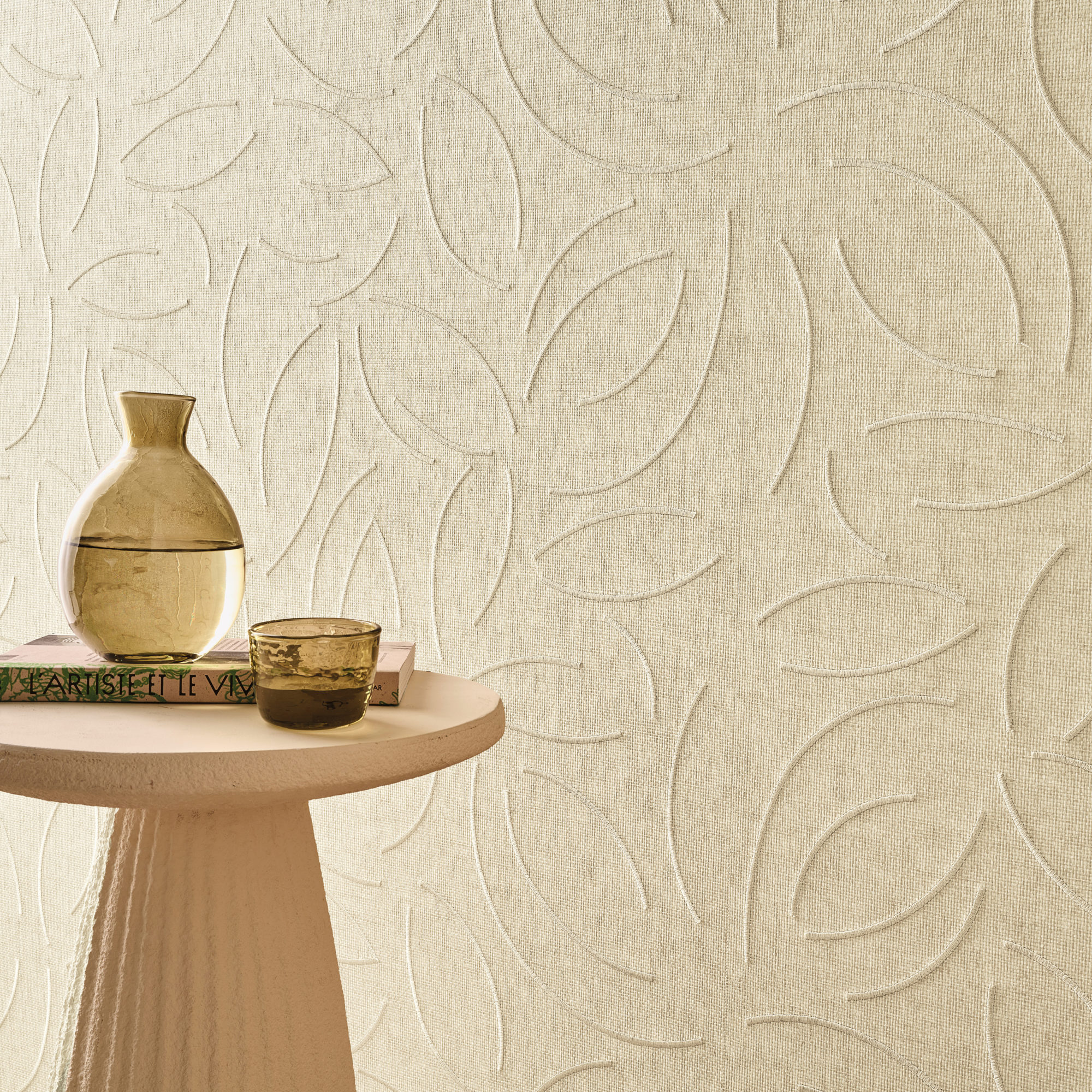 This serene interior highlights Omexco’s OAS221 reference, Verano, from the Oase collection. The wallcovering, crafted from exquisite paperweave, features delicate embroidered patterns inspired by organic forms, adding a sense of movement and sophistication to the space. The soft, neutral tones create an inviting and calming atmosphere, perfectly complemented by a sculptural side table, a glass water carafe, and a textured ceramic cup. The interplay of light and shadow on the embossed surface enhances its depth, making it a refined choice for elegant and natural interiors.