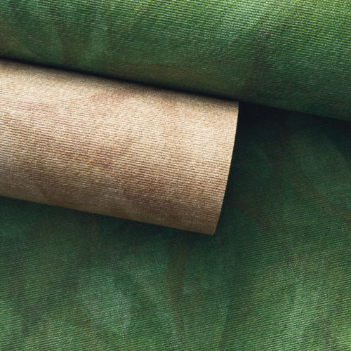 A close-up of two rolls from Omexco’s Oase collection, featuring references OAS201 and OAS203, resting on an unrolled OAS203 wallcovering. The organic patterns and rich green and earthy tones evoke a natural, serene ambiance. The subtle textures and soft color variations highlight the artisanal quality of the collection, inspired by the beauty of nature.