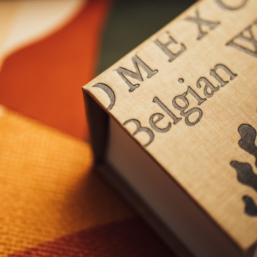 A close-up of a grey Omexco sticky note booklet, containing square sticky notes for quick memos. The booklet rests on the OAS211 reference from the Oase collection, titled Alegre. This panoramic design, inspired by naïve art, evokes the vibrant landscapes of South America through a harmonious blend of warm, earthy tones and organic shapes. The fine texture of the paperweave adds a tactile quality to the composition.