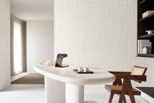 A sophisticated office setting featuring Omexco's Pure collection, reference PU281, a bold wallcovering that showcases a weave of mulberry bark, highlighting the inherent richness and texture of this natural material. The intricate square grid design adds a tactile element and visual depth to the space, emphasizing the organic beauty of the bark. The room includes a sculptural white desk with soft curves, adorned with a modern black desk lamp and a tray holding minimal ceramic cups. To the right, a mid-century inspired wooden chair with cane detailing complements the natural tones of the wallpaper. Built-in shelves displaying books and decor seamlessly integrate with the overall minimalist aesthetic, creating a tranquil and stylish workspace.