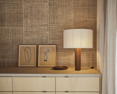 Captivating kitchen closeup featuring Omexco Intuition wallcoverings, meticulously crafted from burlap with an elegant patchwork design and exquisite stitching in a natural brown color. Against the wall, a kitchen cabinet in light wood stands prominently with a stone worktop. On the right side of the image, a linen beige curtain adds a soft touch to the ambiance. On the kitchen cabinet, a wooden table lamp with a white lampshade stands as a stylish accent piece. Additionally, two decorative frames featuring abstract illustrations adorn the cabinet. Two small wooden trays rest atop the cabinet, adding practicality and visual interest to the composition.