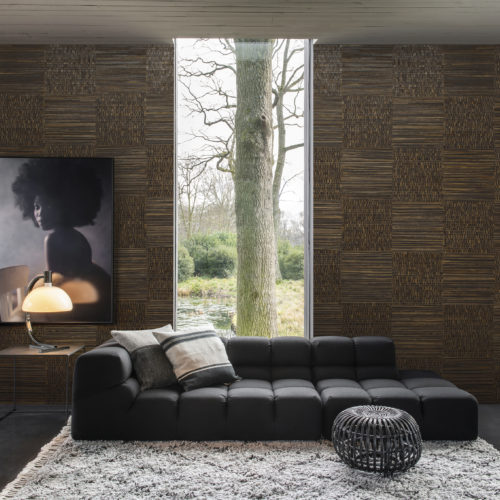 Living room with a anthracite lounge chair, lush carpet and round pouf. On the wall a picture of a girl with an afro. The walls are dressed in woven banana bark: the elegance of this natural material arises from the intricate craftsmanship of an experienced artisan who collects the banana bark from tropical climes. The Omexco Seraya collection.
