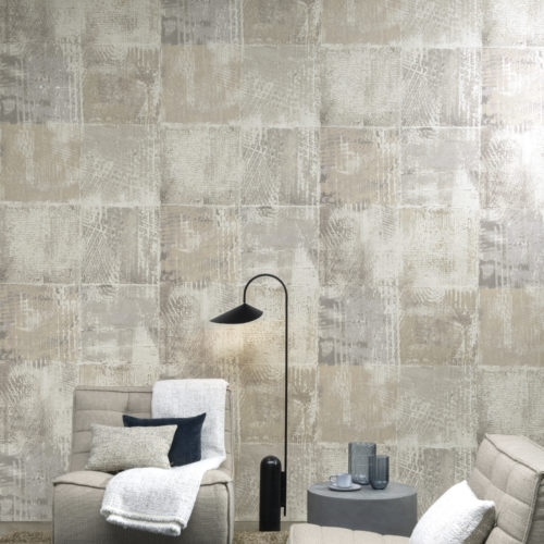 In this image we see a feature wall with organically withered squares printed on a texture of fine opalescent flakes of mica. These wallcoverings are reminiscent of graffitis faded by time. The design is called "Memoirs" and it belongs to the Moonstone collection by Omexco. Two single seater sofas are placed in front of the wall and there is a plush grey carpet on the floor. The shades in this room are neutral: beige, light grey and blue grey tones.