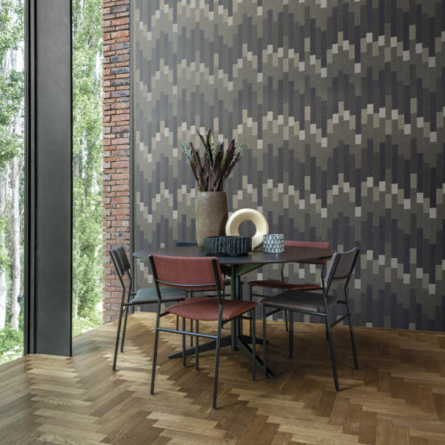 Dining room area with a mosaïc of fine twinkling mica flakes on the wall. The shapes form a rythmic and striking geometric design in shades of dark purple, grey and beige. This is the 'Cadence' design from Omexco's Moonstone collection. We see a dining table with 4 chairs, some decoration on the table, a large window on the left and a wooden herringbone floor.
