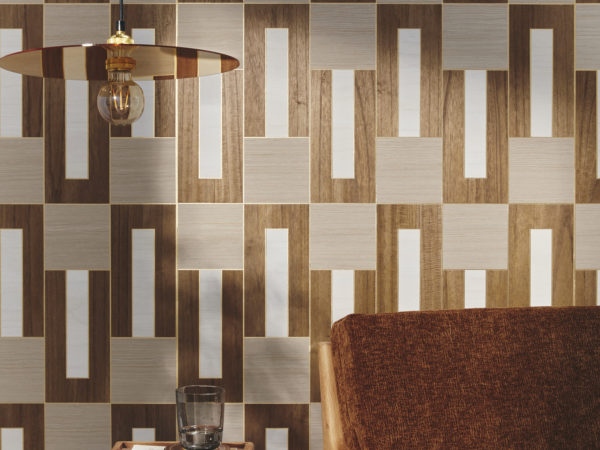 A close-up of a mid-century modern armchair with a wooden side table, adorned with a book and a glass. A sleek, minimalist hanging lamp, showcasing a visible lightbulb, illuminates the scene. Natural daylight gently graces the wall, on which Omexco's marquetry wallcovering collection "Imperialis" is displayed. It showcases a delicate interlocking pattern of wood against a metallic background. The shapes are dark brown, light brown and white, creating a sophisticated and harmonious atmosphere.