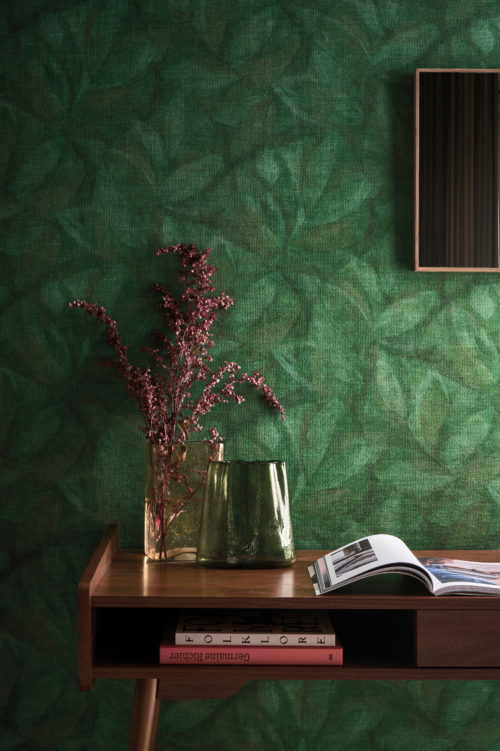 This intimate interior highlights Omexco’s OAS203 reference, Hierbas, from the Oase collection. The wallcovering captures organic beauty in print, depicting delicate layered leaves in deep green tones. The natural texture of the material enhances the botanical motif, creating a soft and inviting ambiance. A sleek wooden console table with warm tones complements the richness of the wall, adorned with minimalist decor elements: a pair of tinted glass vases, a sprig of dried flowers, and a carefully selected book arrangement. A subtle framed artwork completes the setting, reflecting the harmony between nature and design.