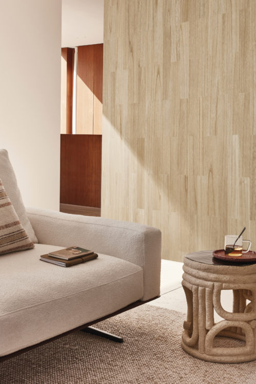 A serene living room corner featuring a beige sofa adorned with a striped pillow and topped with a stack of books. A beige carpet adorns the floor below. Next to the sofa sits an organic-shaped beige round side table, a design by Pascale Degueldre and Jehanne Hupin. The backdrop showcases Omexco's exquisite wood veneer wallcovering collection Imperialis, featuring a parquet-like natural wood design.