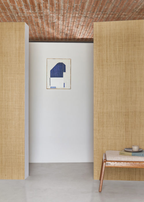 Interior decor featuring a stylish setting with a brick ceiling, grey cast floor, and walls adorned with the Intuition wallcovering collection from Omexco wallcoverings, showcasing a raffia weave in a natural color. On the right, a wooden bench complements the ambiance, adorned with a selection of books.