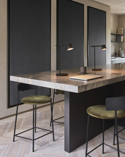 An elegant kitchen scene featuring a luxurious marble countertop adorned with sleek utensils. Two black minimalist table lamps illuminate the space, complemented by three black barstools boasting plush green velvet seating. In the background, three panels showcase Omexco's acoustic wallcovering collection, Silence, in the sophisticated color 'anthracite,' adding a touch of modernity and acoustic functionality to the chic kitchen ambiance.
