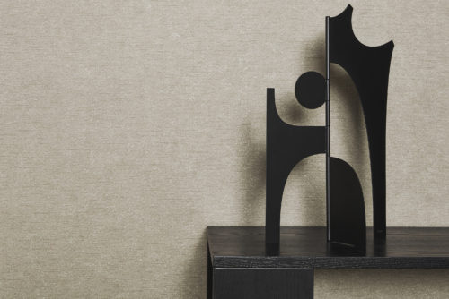 A captivating close-up of a black wooden table adorned with a minimalist organic sculpture. The backdrop features Omexco's acoustic wallcoverings, Silence, in a soothing beige tone, offering a soft and tactile feel. These wallcoverings not only contribute to the aesthetic appeal but also possess the remarkable ability to enhance acoustic comfort.