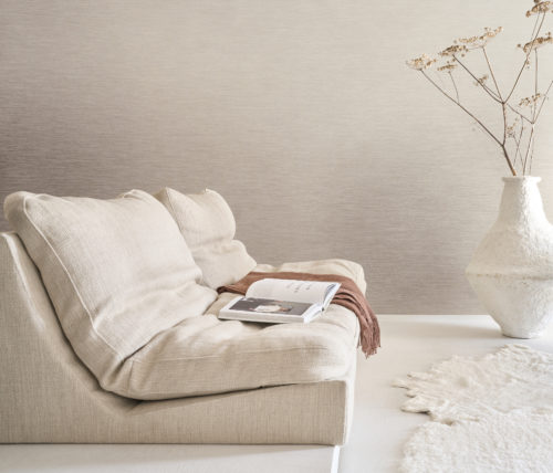 A serene living room featuring Omexco's Pure collection, reference PU351, composed of twisted textile warps crafted from noble materials such as linen. The wallcovering’s refined texture adds subtle depth and sophistication, creating an understated yet elegant atmosphere. The scene includes a low, plush sofa in soft beige tones, styled with a draped rust-colored throw and an open book. To the right, a large, sculptural white vase holds delicate dried flowers, while a soft, irregularly shaped white rug enhances the minimalist aesthetic of the space. The overall design harmoniously combines natural materials and muted tones to evoke a tranquil, refined interior.