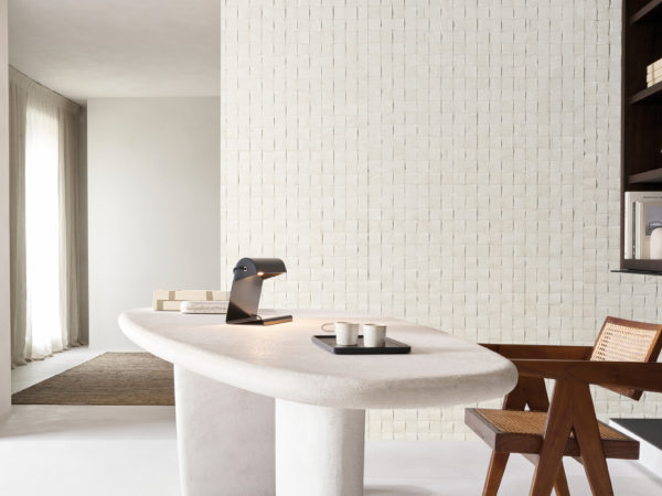 A sophisticated office setting featuring Omexco's Pure collection, reference PU281, a bold wallcovering that showcases a weave of mulberry bark, highlighting the inherent richness and texture of this natural material. The intricate square grid design adds a tactile element and visual depth to the space, emphasizing the organic beauty of the bark. The room includes a sculptural white desk with soft curves, adorned with a modern black desk lamp and a tray holding minimal ceramic cups. To the right, a mid-century inspired wooden chair with cane detailing complements the natural tones of the wallpaper. Built-in shelves displaying books and decor seamlessly integrate with the overall minimalist aesthetic, creating a tranquil and stylish workspace.