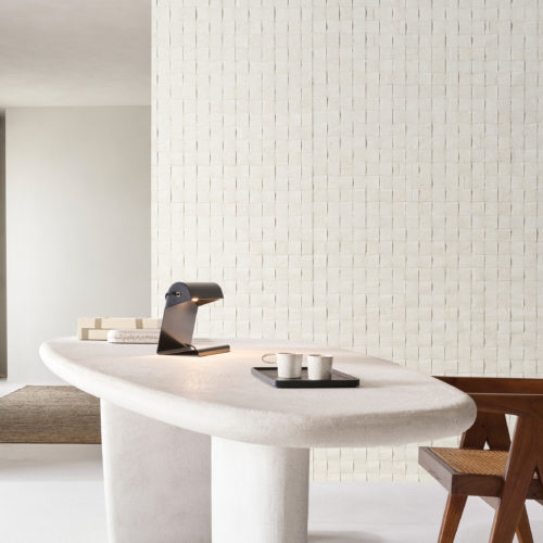 A sophisticated office setting featuring Omexco's Pure collection, reference PU281, a bold wallcovering that showcases a weave of mulberry bark, highlighting the inherent richness and texture of this natural material. The intricate square grid design adds a tactile element and visual depth to the space, emphasizing the organic beauty of the bark. The room includes a sculptural white desk with soft curves, adorned with a modern black desk lamp and a tray holding minimal ceramic cups. To the right, a mid-century inspired wooden chair with cane detailing complements the natural tones of the wallpaper. Built-in shelves displaying books and decor seamlessly integrate with the overall minimalist aesthetic, creating a tranquil and stylish workspace.