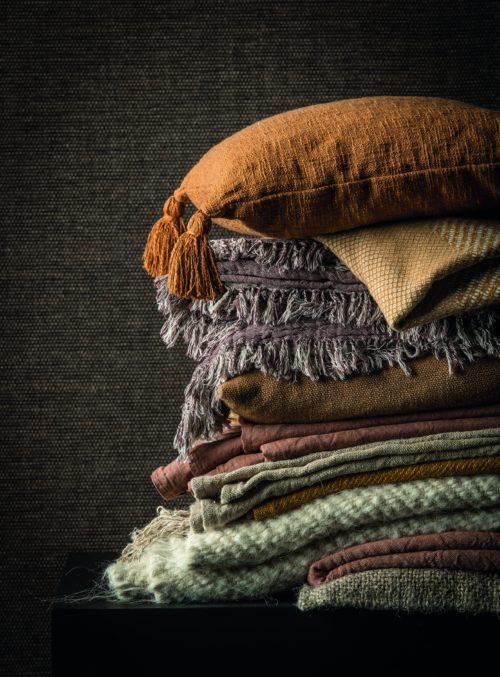 L'uni subtil et serein de la collection de tissage de papier Tribu d'Omexco. Nous voyons un gros plan du motif Duo, un tissage magnifiquement simple dans des tons anthracite et chocolat. C'est la toile de fond idéale pour les coussins et les couvertures colorés que l'on voit au premier plan de cette photo.