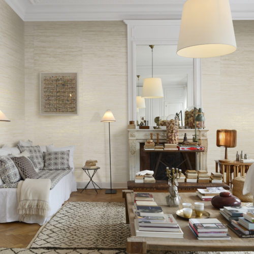 Nous voyons un grand salon. Les canapés sont tous enveloppés dans des draps blancs. Il y a une grande table basse avec beaucoup de livres. Cette pièce chaleureuse est parfaite pour nos fibres de raphia tissées : des fibres de raphia tissées sur des supports dorés, argentés ou cuivrés pour refléter la lumière. Après le tissage, l'artisan enduit les fibres de tons métalliques.