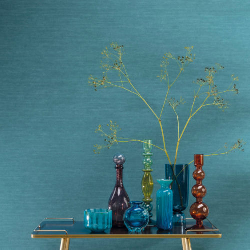 Gros plan sur un mur turquoise vif. Il s'agit de l'uni le plus populaire d'Omexco, imprimé sur un intissé texturé pour lui donner un aspect textile familier. Le motif de ce revêtement mural s'appelle "Silky" et appartient à la collection tropicale "Birds of Paradise". Devant le mur, nous voyons une table d'appoint dorée sur laquelle sont posés des vases en verre colorés de différentes hauteurs et formes.