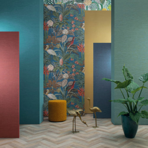 Espace intéressant avec un sol clair à chevrons et six panneaux colorés de différentes hauteurs, allant de la mi-hauteur à la hauteur du plafond. Au sol, on trouve un pouf rond ocre, une grande plante d'intérieur tropicale dans un pot en céramique vert et deux statues dorées de grues. Les panneaux sont décorés de revêtements muraux unis de couleur jaune, turquoise et bleu foncé, ainsi que de motifs tropicaux représentant de fabuleuses fleurs, des feuilles luxuriantes et de magnifiques oiseaux. La collection de revêtements muraux "Birds of Paradise" d'Omexco est illustrée ici.