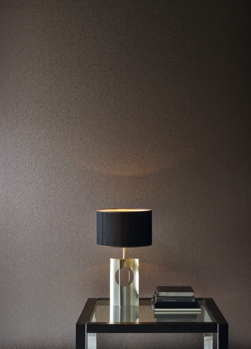 Mur impressionnant avec le revêtement mica de la gamme Omexco Graphite. Table en verre avec détails noirs, abat-jour noir mat et quelques livres.