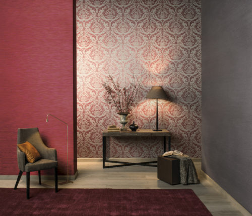 Grande pièce aux couleurs rouge, violet et gris. On y trouve un fauteuil en cuir, un tabouret avec une couverture douce, un livre, une table avec des branches de baies. Le tout combiné avec le revêtement mural Omexco Elegance uni et damassé.