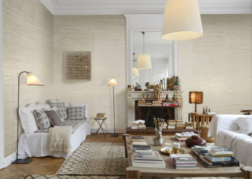 Nous voyons un grand salon. Les canapés sont tous enveloppés dans des draps blancs. Il y a une grande table basse avec beaucoup de livres. Cette pièce chaleureuse est parfaite pour nos fibres de raphia tissées : des fibres de raphia tissées sur des supports dorés, argentés ou cuivrés pour refléter la lumière. Après le tissage, l'artisan enduit les fibres de tons métalliques.