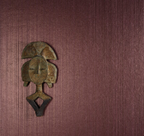 Mur avec un revêtement mural bordeaux, reflétant la lumière. Un masque africain en bois nous regarde.