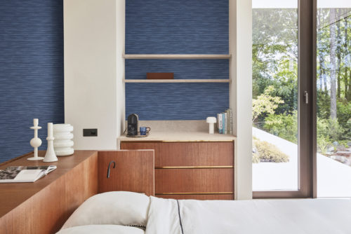 Une chambre à coucher contemporaine à l'ambiance chaleureuse, dotée d'une tête de lit en bois, d'un élégant meuble en bois à tiroirs et d'étagères murales encastrées. Sur la droite, une porte vitrée s'ouvre sur un jardin luxuriant. Les murs présentent la collection haute performance Rivage d'Omexco en bleu lapis, avec une structure subtile et délicatement surélevée qui rappelle le contour serein d'un horizon lointain, ajoutant une touche de sophistication à l'espace moderne.